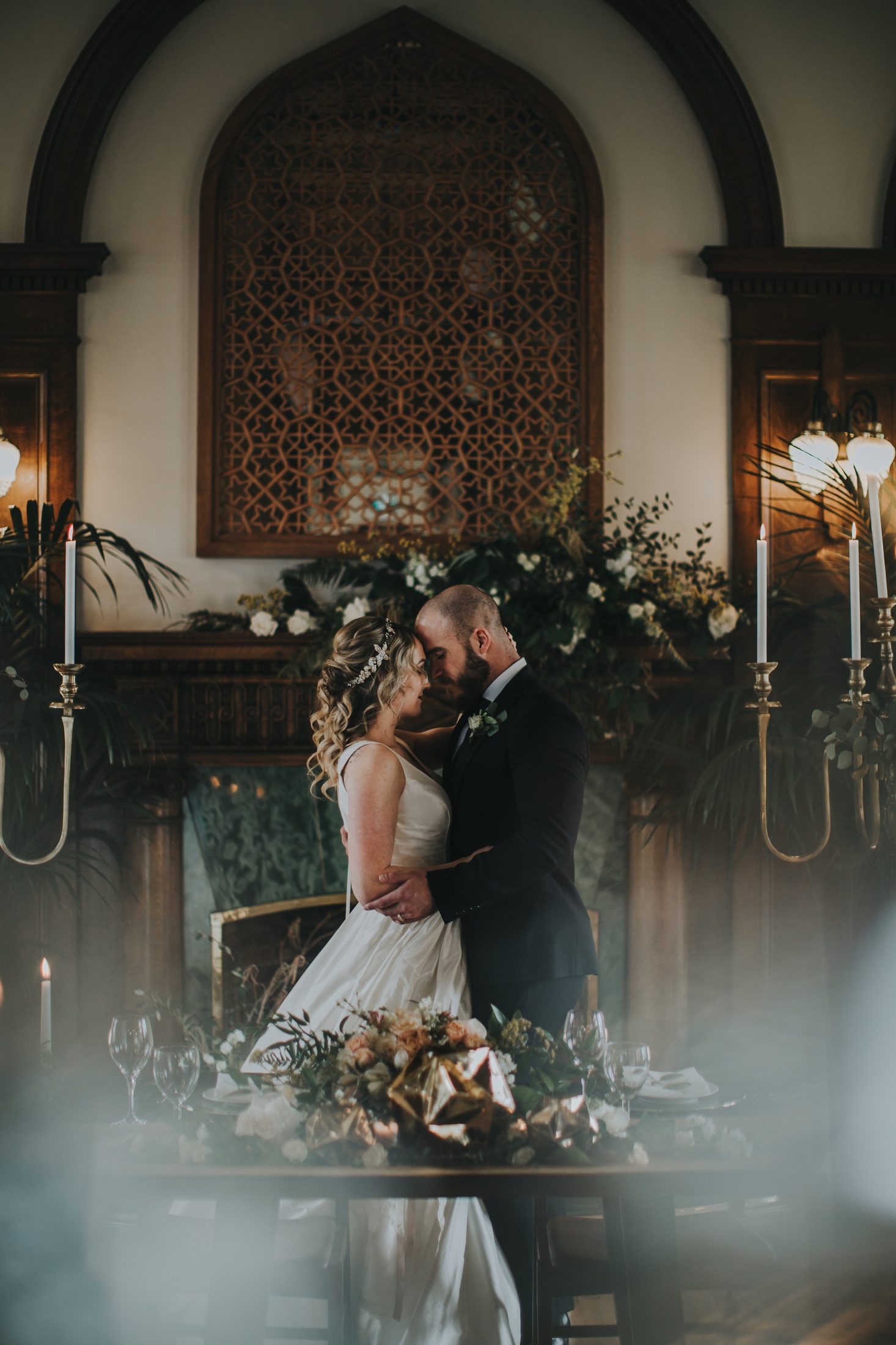 The couple beside an ornate fireplace mantel, styled with candelabras and floral details.